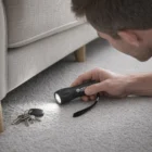 A man uses a Modo Torches flashlight to look for keys under a gray couch on carpet.