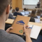 Person taking notes in a lecture hall, using a Green Eco Stone Paper Pad labeled Pixel Cloud Data Maps.