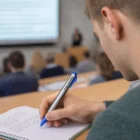 Student taking notes with a Lamy Vivo Ball Pen in a lecture hall, blurred screen behind.