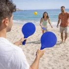 Three people play paddle ball with Racket And Ball Sets on a sandy beach by the ocean.