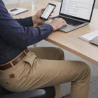 Man in Gents Boston Chino Pants at desk using laptop and phone, checking emails at work.