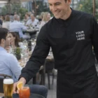 Smiling waiter in Mens Long Sleeve Continental Shirt serves drinks outdoors.