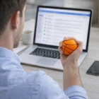 Man at laptop with emails open squeezes a Stress Balls BasketBalls orange stress toy.