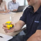 Person holding a yellow Promo Stress Balls Beatle Car in a meeting room with others in background.