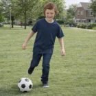 Smiling child in Kids Cut Crew Ringspun Neck Tshirt plays soccer on sunny park grass.