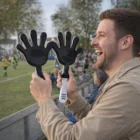 Man smiles holding two large black The Happy Hand Klappers at an outdoor sports game.