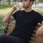 Man in a black Mens Stretch Fit Personalized T Shirt drinks from a cup on a park bench.