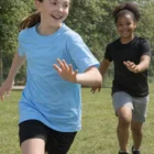 Two girls in Cool Dry Customised Kids T Shirts run and smile outdoors on a sunny grassy field.