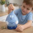 Smiling boy puts a coin into Wally The Water Drop Coin Savings Bank on the table.