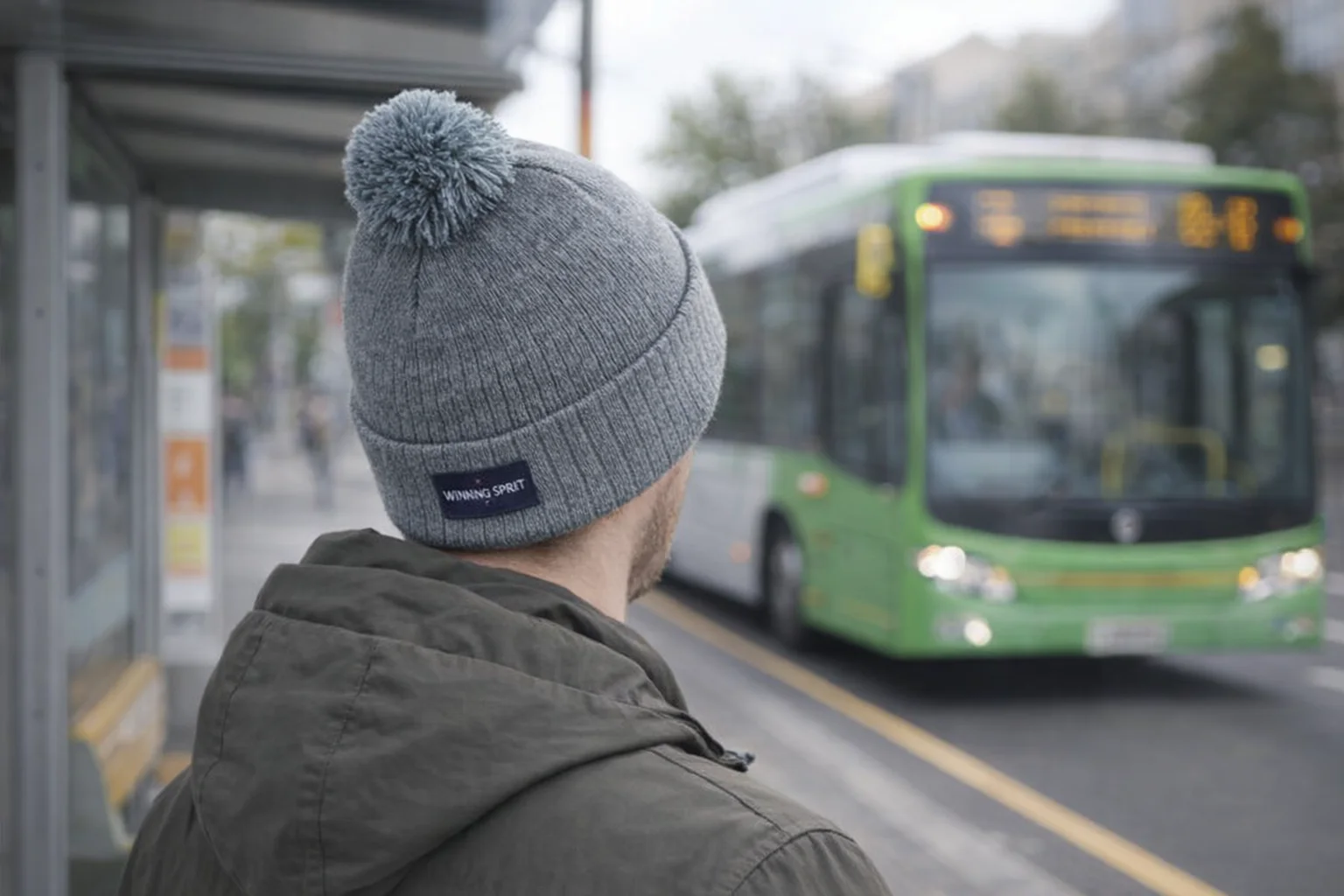 Person in an Acrylic Beanies With Pompon waits at a bus stop as a green bus approaches.