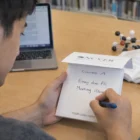 Person writing a to-do list with Sticky Notes Combo A beside a laptop and books on the table.
