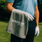Golfer in blue shirt holds a gray VADEN ORIGINAL towel draped over their arm.