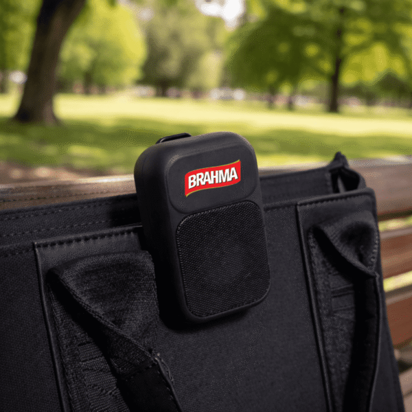A black BRAHMA speaker clipped to a black bag on a park bench, with trees in the background.