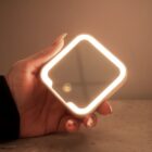 A hand holds a small, square lighted mirror with a glowing frame on a gray surface.