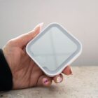 A hand holds a small, white, square device with a reflective surface.