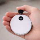 A hand holding a round white device with a black button labeled YES on top.