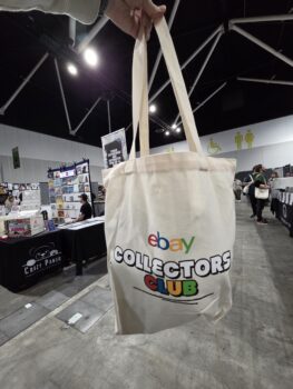 A hand holds an eBay Collectors Club tote bag at an indoor convention or expo.