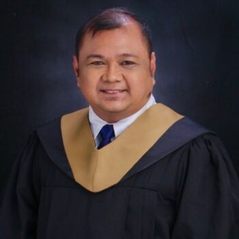 Meet the team: Smiling person in graduation gown with gold and black collar, dark background.