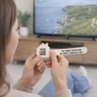 Person holding House Shape Branded Emery Boards, seated by a TV with a coastal scene.