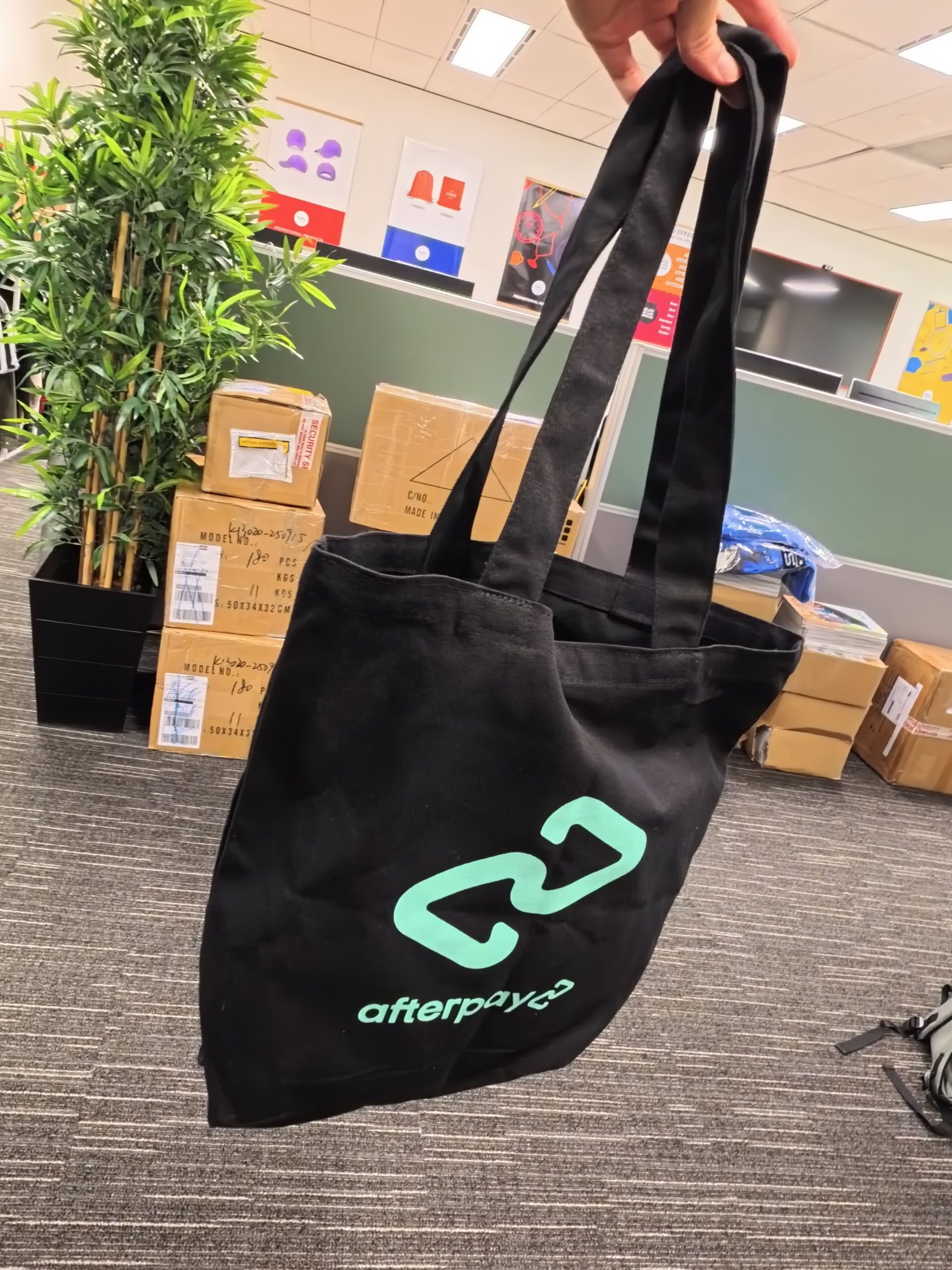 A hand holds a black tote bag with a green 23 afterpay logo—perfect for 5 items for events.