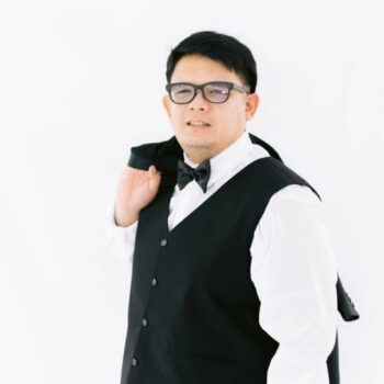 Meet the team: Man in a black vest and bow tie, holding his jacket over his shoulder.