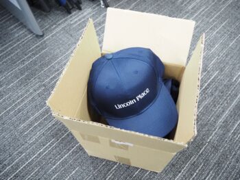 A blue Lincoln Place cap in an open cardboard box on a gray carpet.