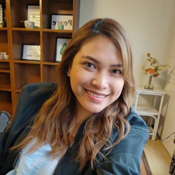 Smiling woman with long hair sits indoors, meet the team amid wooden shelves and flowers.