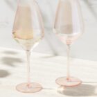 Two pink-tinted wine glasses, one with white wine, on a light surface with shadows.