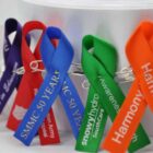 Colorful awareness ribbons with printed text pinned to a white background using safety pins.
