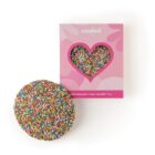 Large chocolate disc with colorful candy sprinkles beside a pink heart-shaped windowed box.