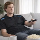 Man in a Gildan Hammer Adult T-Shirt sits on a couch with popcorn and a mug nearby.