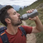 Man in red shirt drinks from Large Super Branded Bottle while hiking a mountain trail.