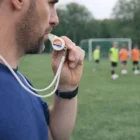 A coach uses a Wavo Sports Whistle while watching kids play soccer on the grass field.