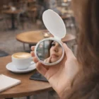 A woman’s face reflected in a Printed Compact Mirror at a café, coffee on the table.