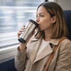 Woman in a beige coat sips from a Basic Budget Thermal Mug while sitting on a train.