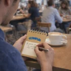 Person writing in Printed Eco A7 Note Pads at a café, coffee cup beside the notepad.