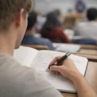 Student takes notes in a lecture hall using Promotional Big Grip Pens; others study nearby.