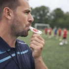 Man blows a Shrill Regular Whistle on a soccer field, players blurred in background.