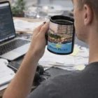 Man with Full Colour Printed Black Mug at messy desk with laptop, papers, and hotel mug.