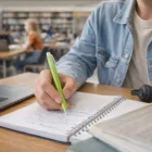 Student writing notes with Light Green Kermit Pens at a library table near a laptop.