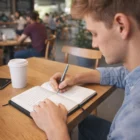 Young man writing in a Pu Leather Medium Notebook at a café table with coffee.