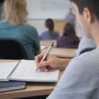 Student in class writing notes with a Crios Pen, surrounded by peers and a teacher in front.