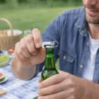 Someone uses The Big Gulp Bottle Openers to open a green bottle at a picnic.