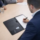 Person in a suit writes on an A4 Corporate Branded Portfolio at a meeting table.