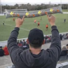 Fan holding a large Trubble Supporters Sticks inflatable at a sports game, field in background.
