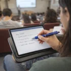 Student taking notes with Muxi Stylus Pens Classic on a tablet in a crowded lecture hall.