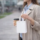 Woman in a beige coat holds Small Printed Paper Bags and a takeaway coffee cup outdoors.