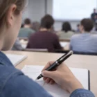 Student taking notes with Nautica Rolling Ball Pens in class, others in the background.