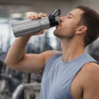 Man at gym drinks from 750Ml Flip Cap Aluminium Bottle Branded, wearing a sleeveless shirt.