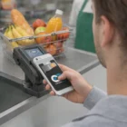 A customer pays at checkout with a Comet Phone Wallet; fruit basket visible in the background.